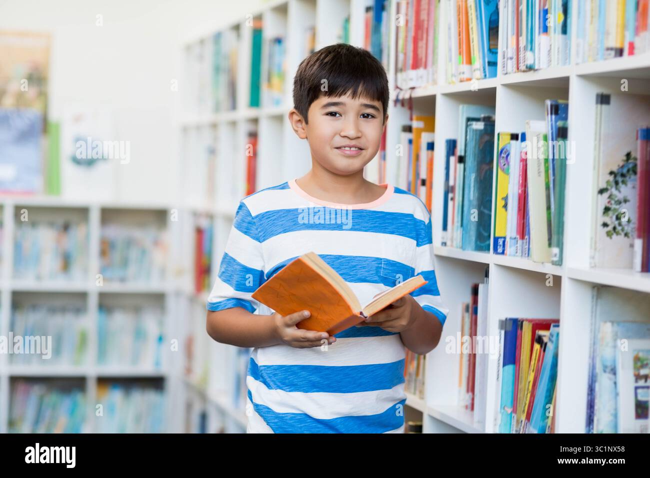 Garçon asiatique lisant un livre orange tout en se tenant dans l'allée de la bibliothèque entre les étagères blanches Banque D'Images