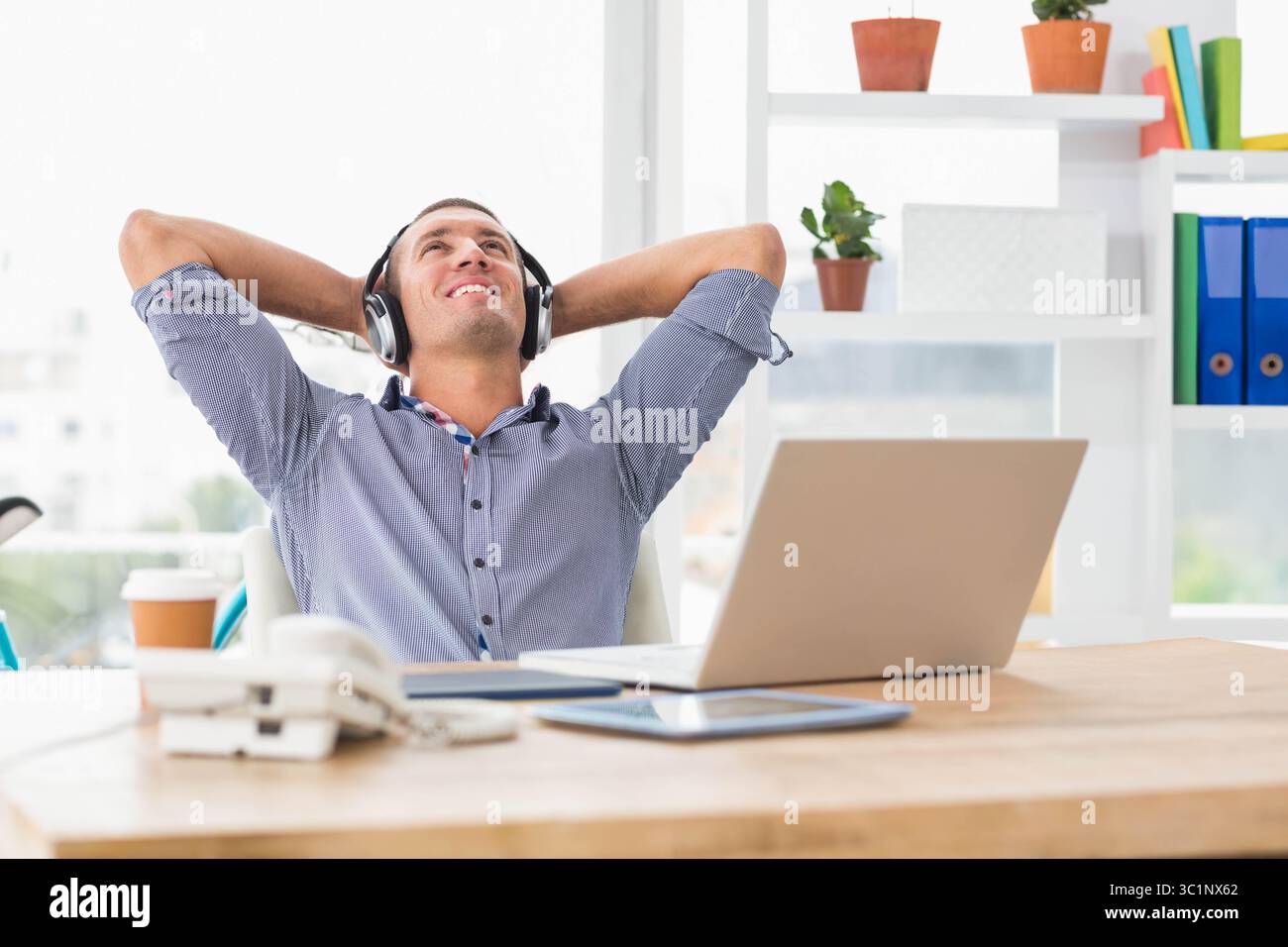 Homme penché au bureau portant des écouteurs avec ordinateur portable, téléphone, café, plantes, classeurs sous la vue de la ville Banque D'Images