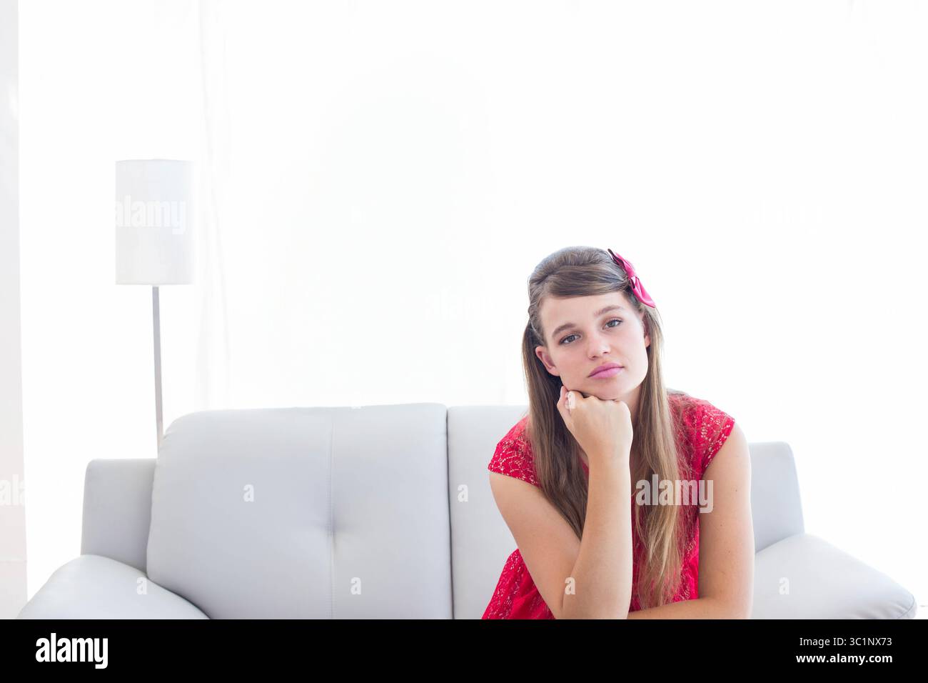 Femme assise sur le canapé portant une robe rouge par lampadaire avec abat-jour blanc, rideaux, espace de copie Banque D'Images
