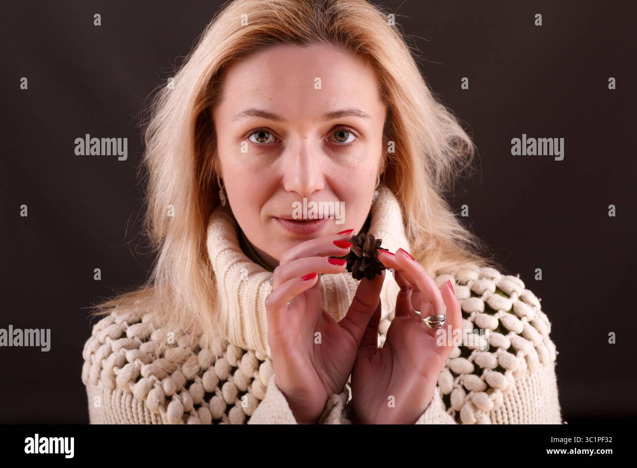 Une femme à la peau claire aux cheveux blonds et aux yeux bleus tient une pomme de pin dans ses mains. Elle porte un pull en tricot texturé de couleur crème et un s. Banque D'Images