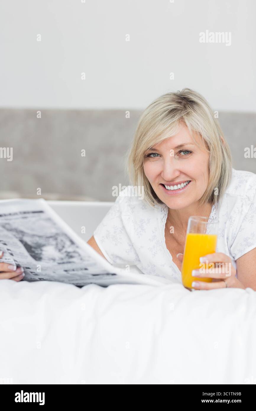 Femme allongée sur le lit dans la chambre tenant un verre de jus d'orange et lisant le journal, espace de copie Banque D'Images