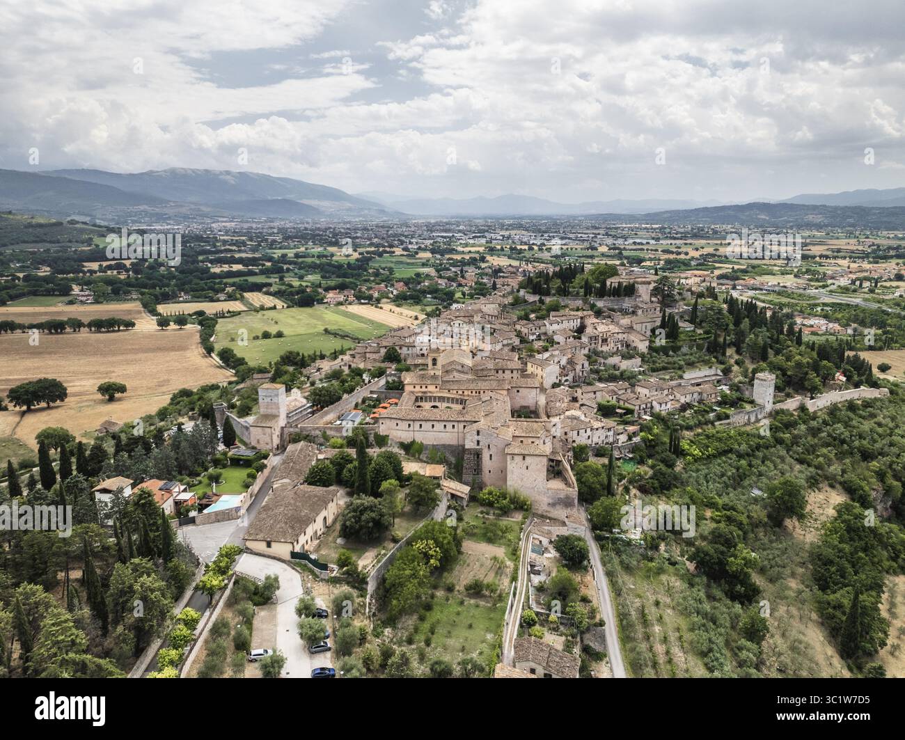 Vue aérienne de bâtiments en pierre baignés de soleil nichés dans des champs verdoyants et des collines ondulantes sous un ciel nuageux, Spello, Ombrie, Italie. Banque D'Images
