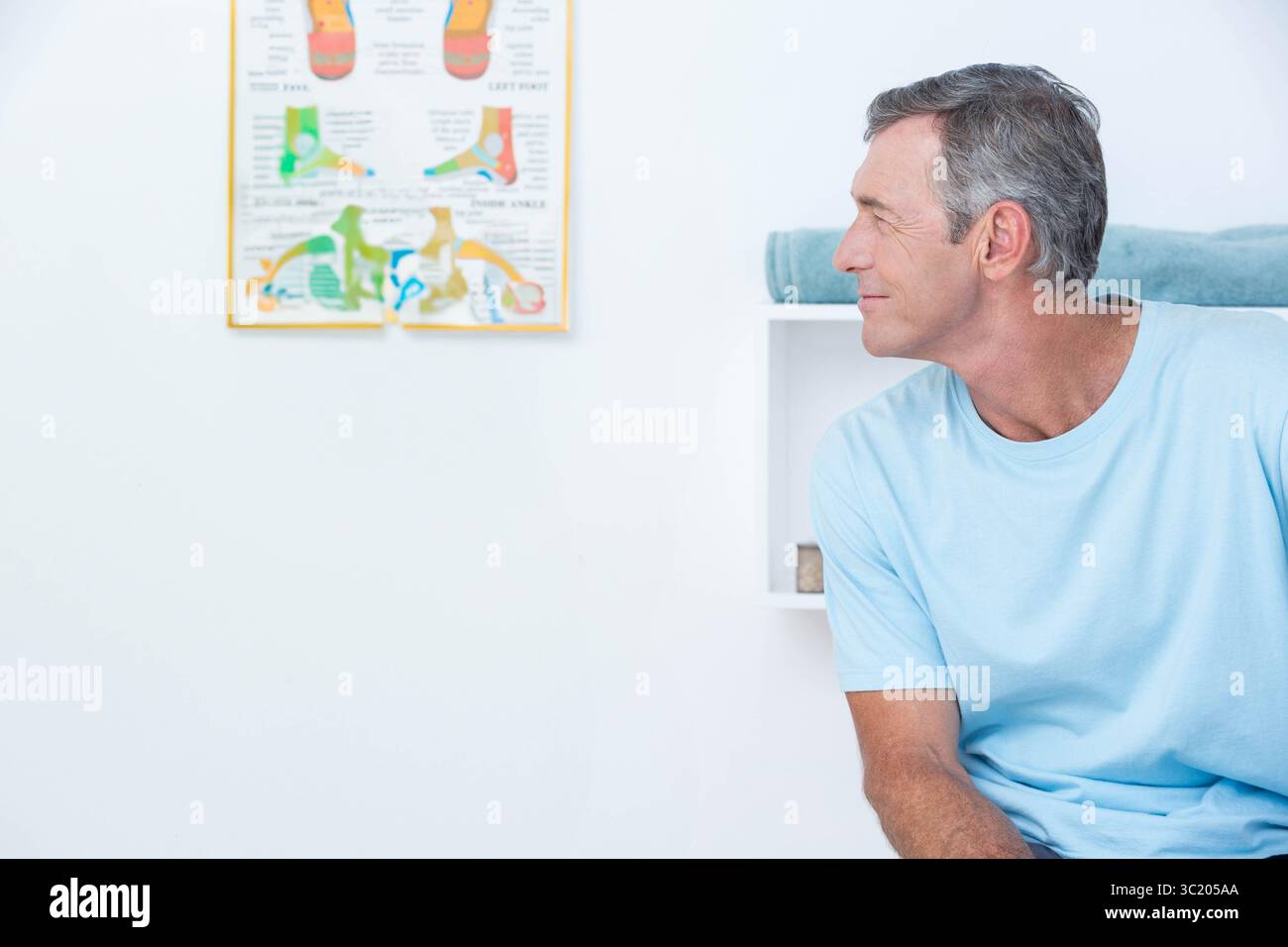 Homme mûr assis sur la table rembourrée dans le cabinet médical regardant la carte anatomique du pied, espace de copie Banque D'Images