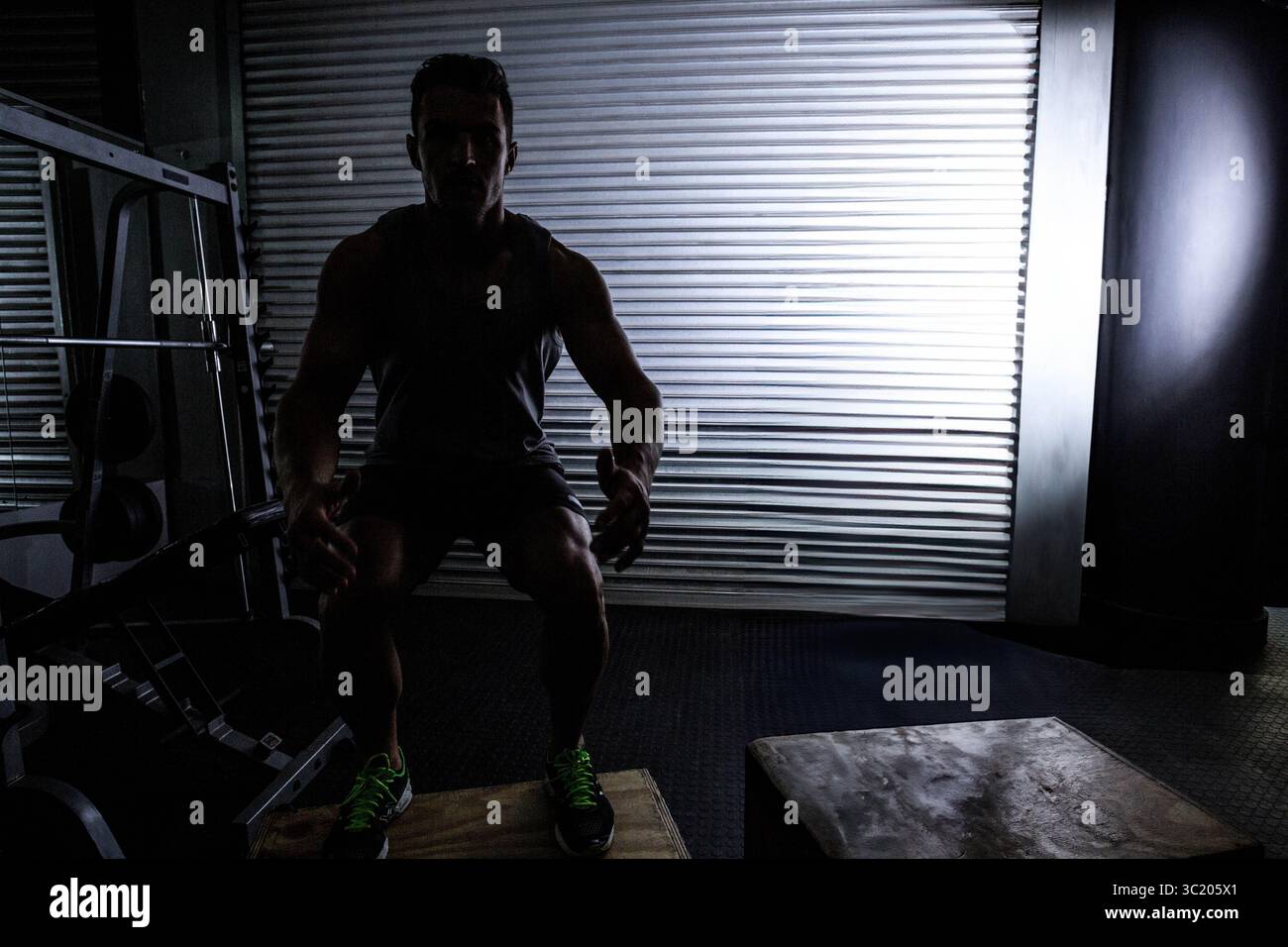 Athlète squattant sur une boîte en bois plyo dans une salle de gym faiblement éclairée avec support de squat et sac de punching Banque D'Images