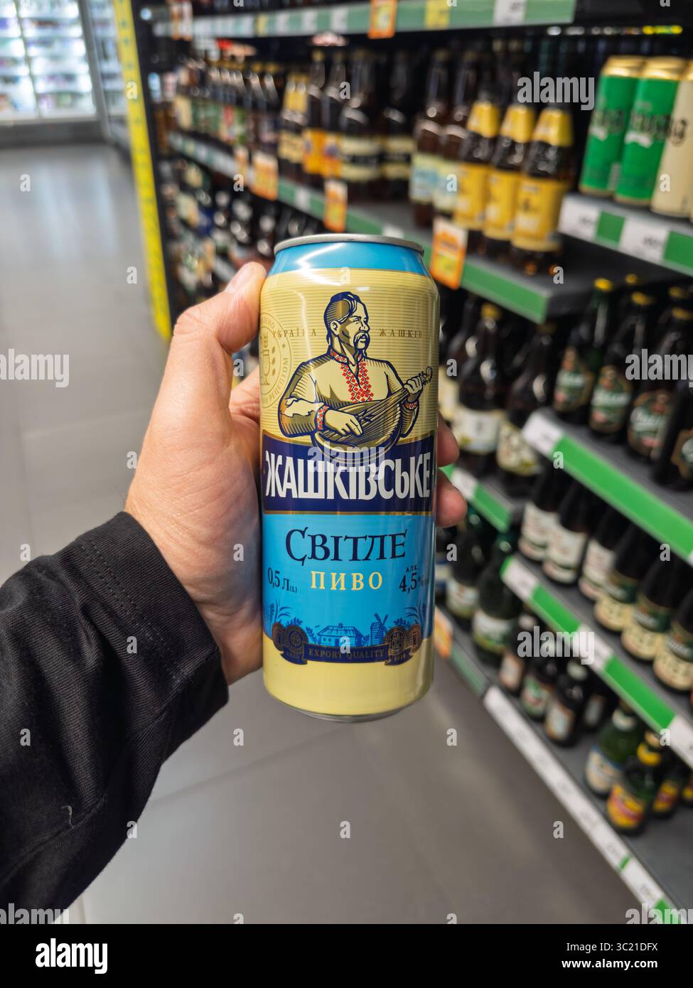 Kiev, Ukraine - 05 juillet 2025 : une main tenant une canette de bière légère ukrainienne Zhashkivske Svitle dans une allée de bière de supermarché Banque D'Images