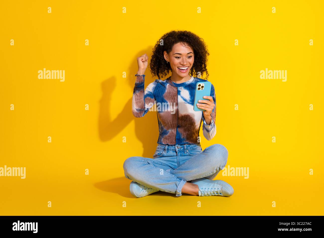 Femme dans le haut vibrant tie-dye célèbre tout en utilisant un smartphone, sur un fond jaune audacieux, exsudant la joie et le style Banque D'Images