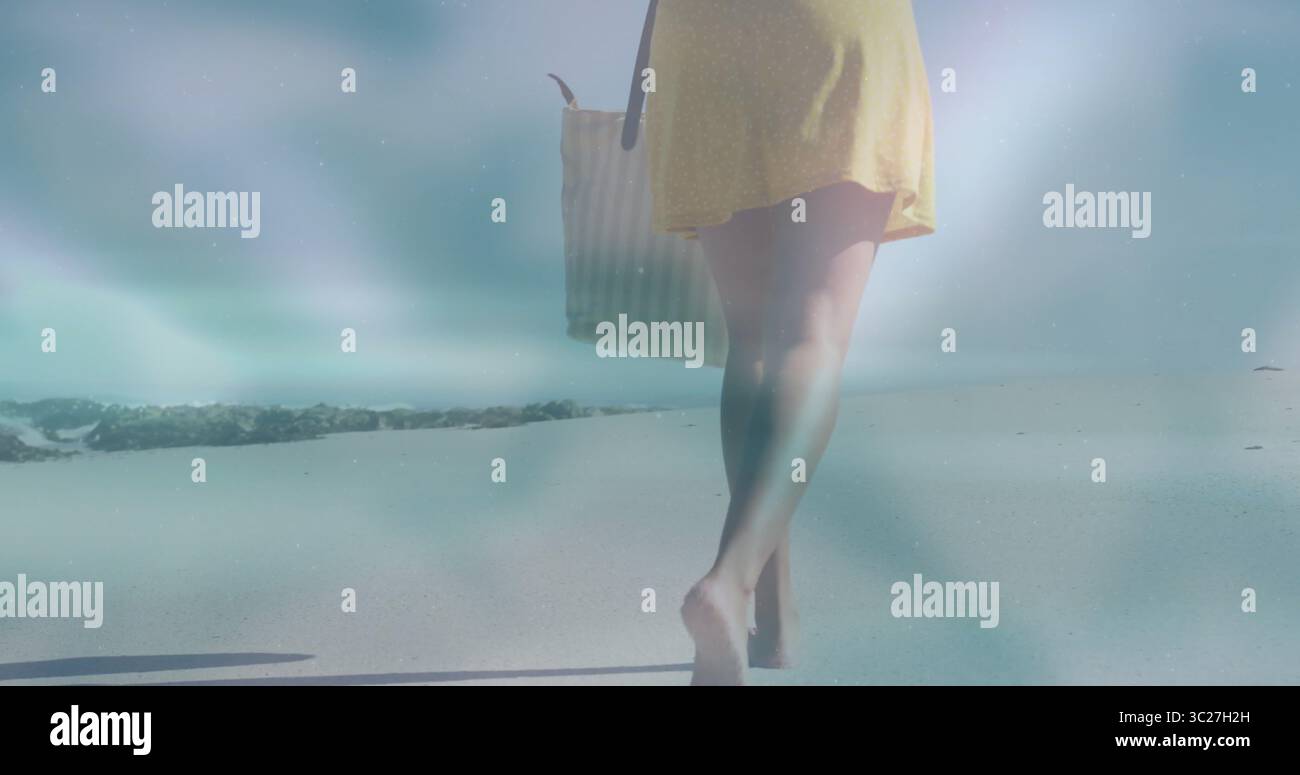 Femme marchant portant une robe courte jaune le long de la plage de sable par brise-lames, portant sac fourre-tout rayé Banque D'Images