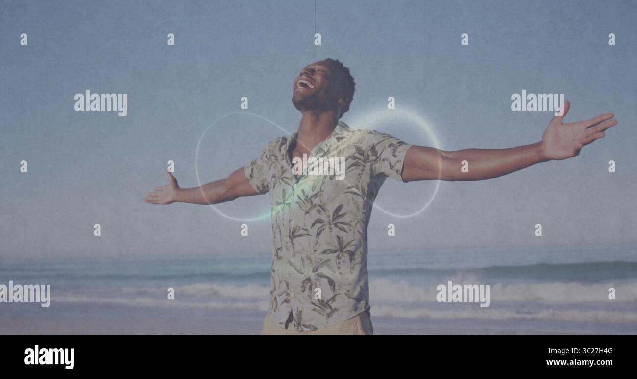 Homme écartant les bras portant une chemise à manches courtes et imprimé palmiers souriant au rivage sablonneux avec les vagues de l'océan Banque D'Images