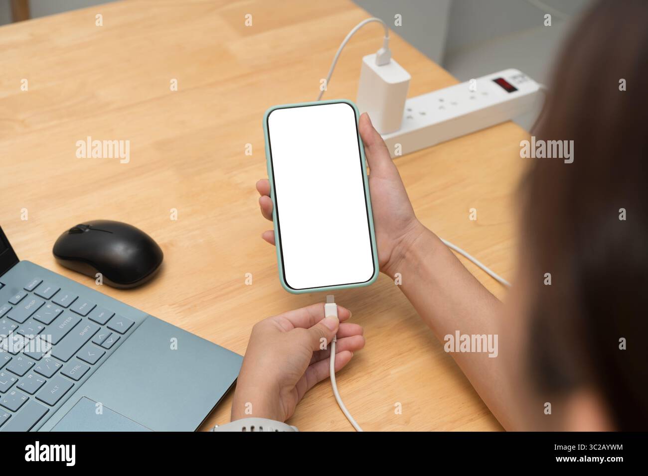 Une personne connecte un smartphone à un chargeur tout en utilisant un ordinateur portable sur une table en bois. Écran vide sur l'appareil mobile. Concept de multitâche, espace de travail, Banque D'Images
