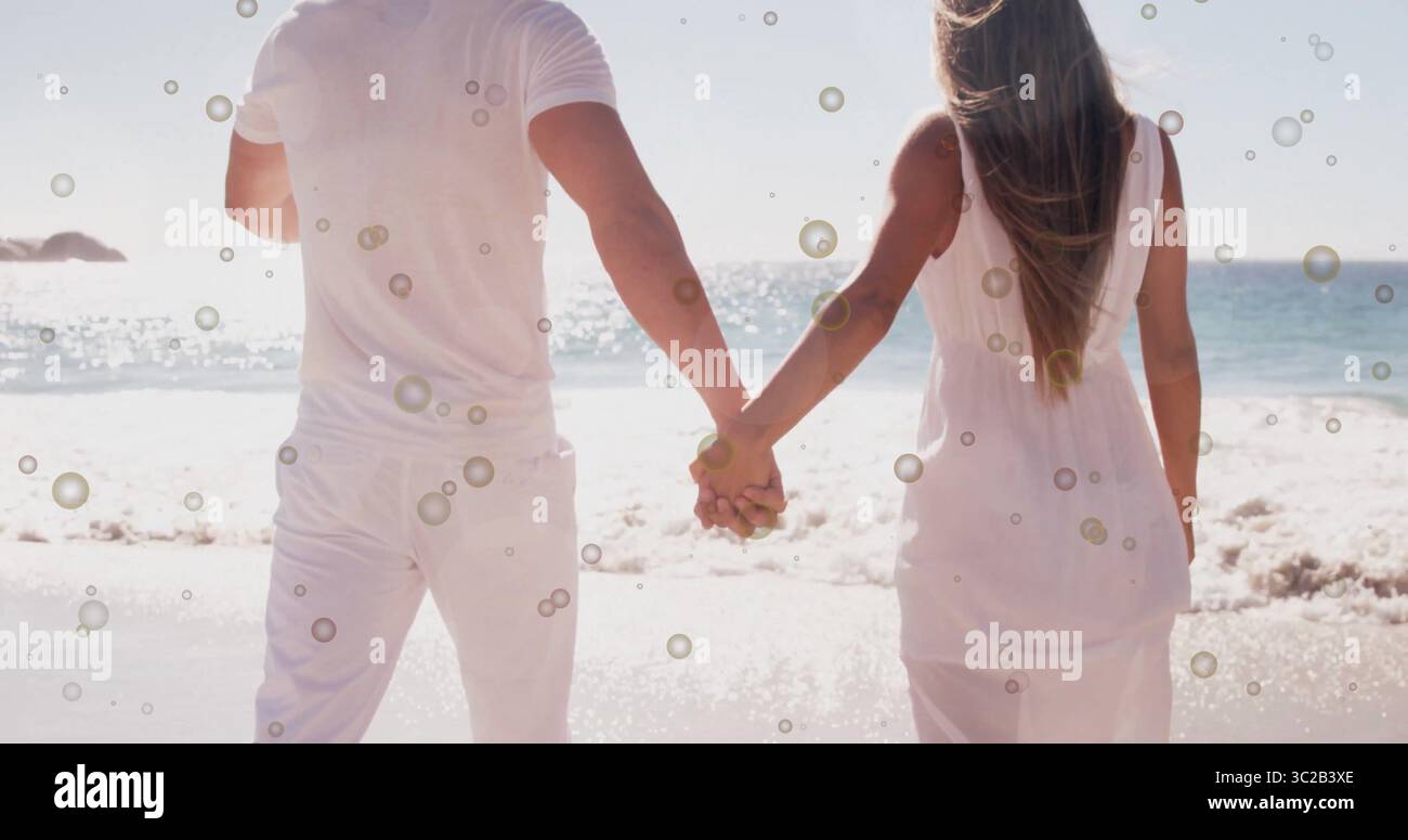 Tenant la main homme et femme à la peau claire dans des tenues blanches debout sur la plage de sable, regardant l'océan Banque D'Images