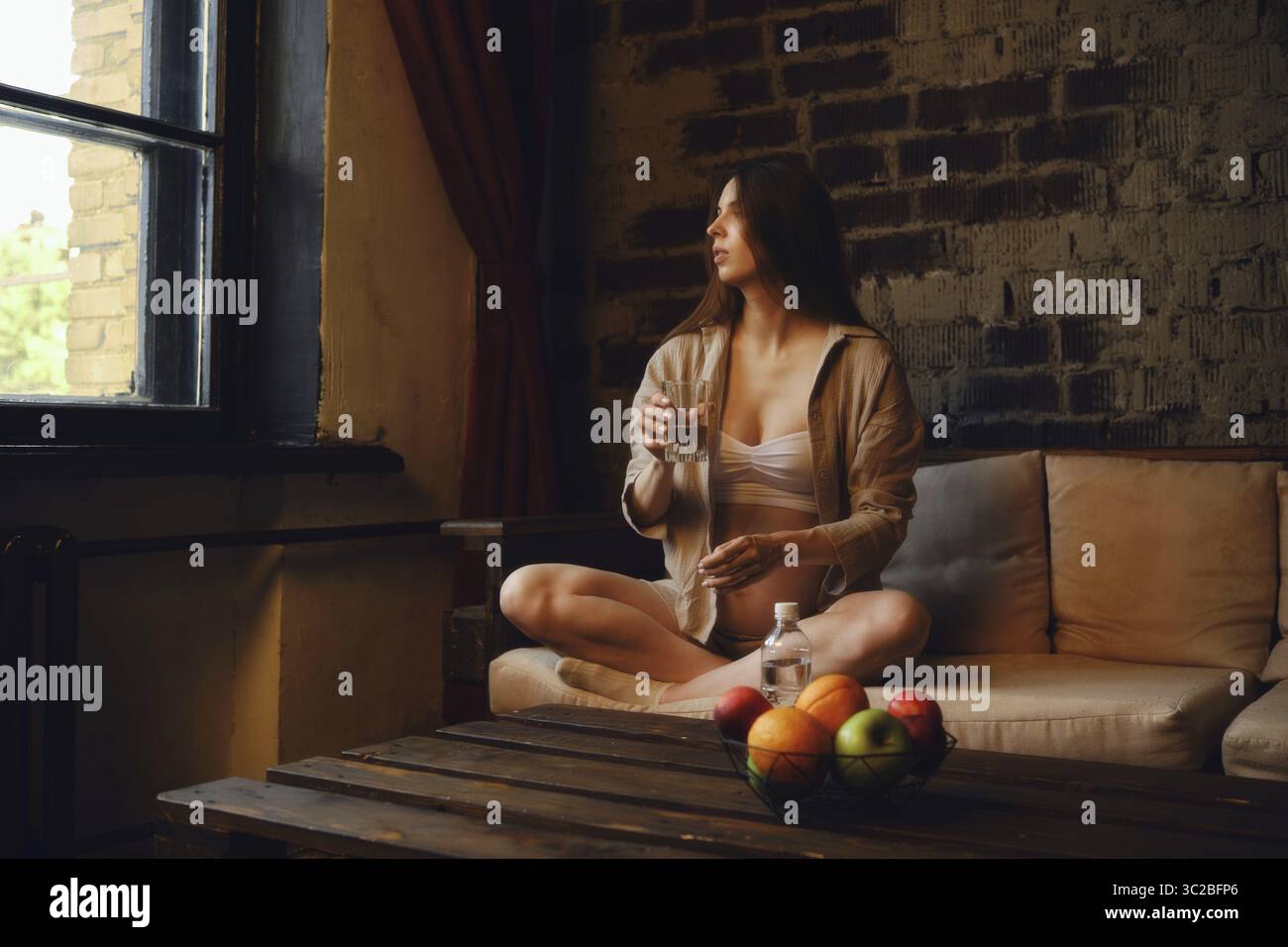 Dans un cadre confortable, une femme enceinte est assise confortablement sur un canapé avec un verre d'eau minérale, regardant par la fenêtre Banque D'Images