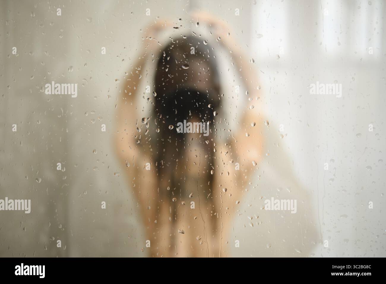 Une femme se tient sous la douche avec les cheveux baissés, profitant d'un moment de paix. L'eau coule le long de son corps, créant une atmosphère sereine avec des gouttelettes Banque D'Images