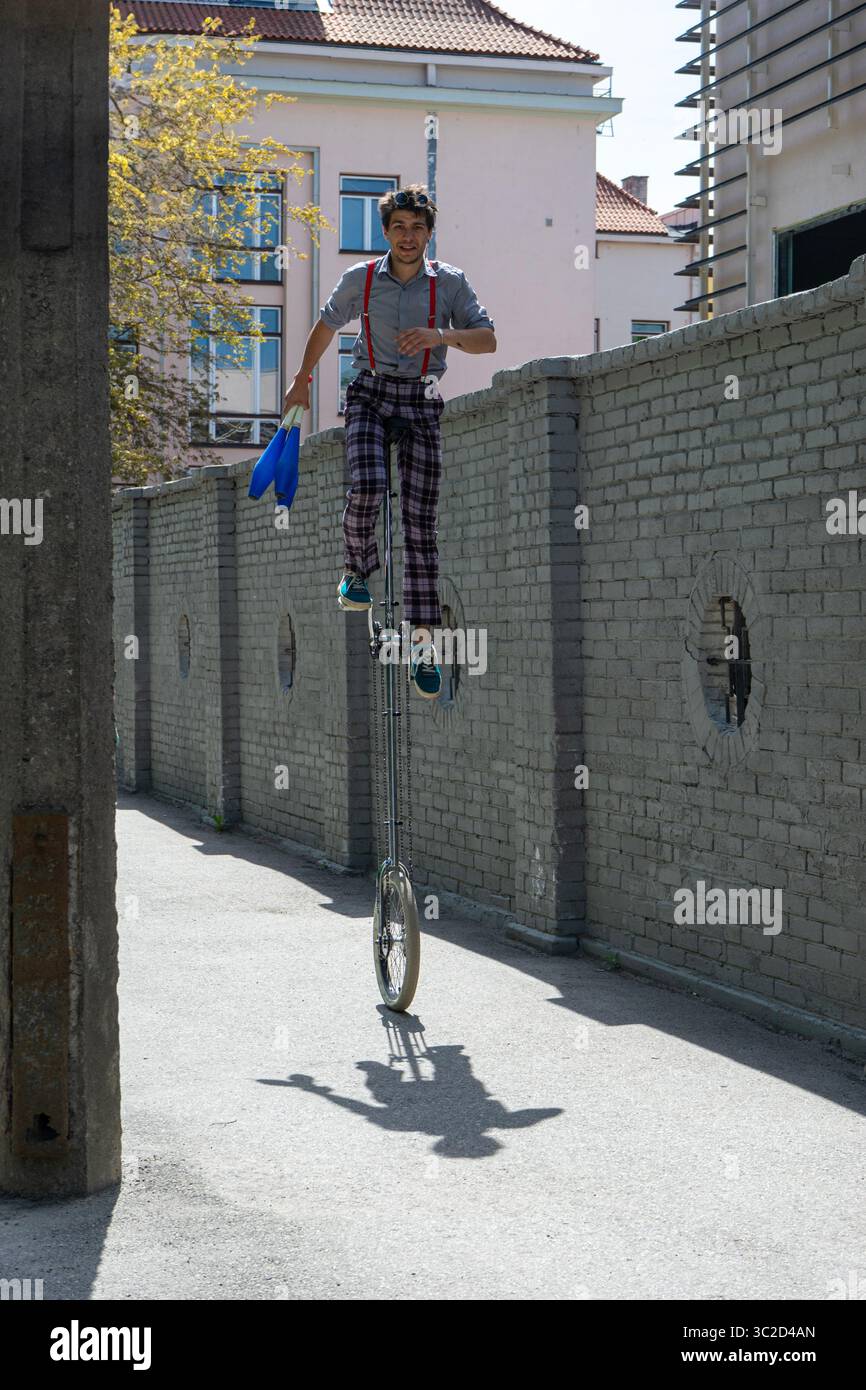 Un artiste de rue chevauchant habilement un grand monocycle dans un cadre urbain, vêtu d'une tenue décontractée avec un chapeau et des bretelles. Banque D'Images