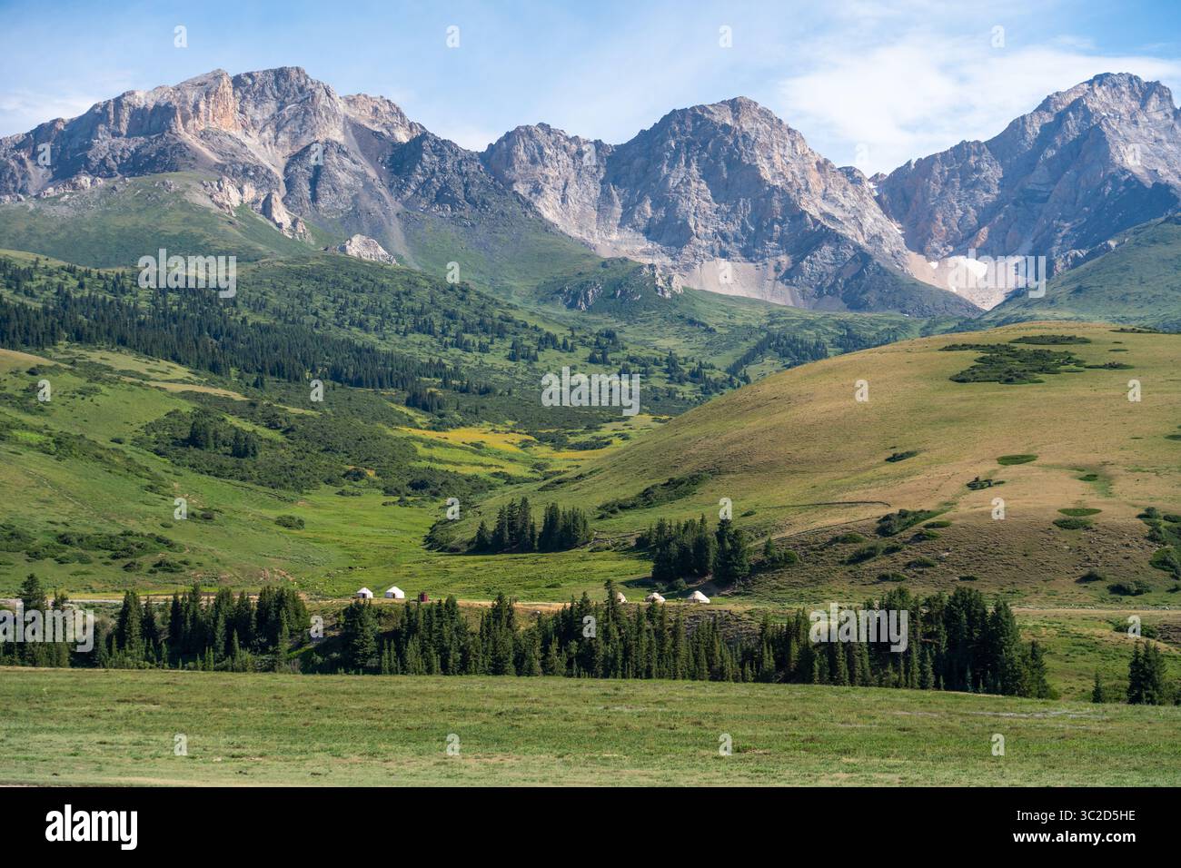 Une vallée de montagne pittoresque avec une ligne de yourtes nichée au milieu de forêts verdoyantes et de collines ondulantes, avec des pics accidentés en arrière-plan Banque D'Images