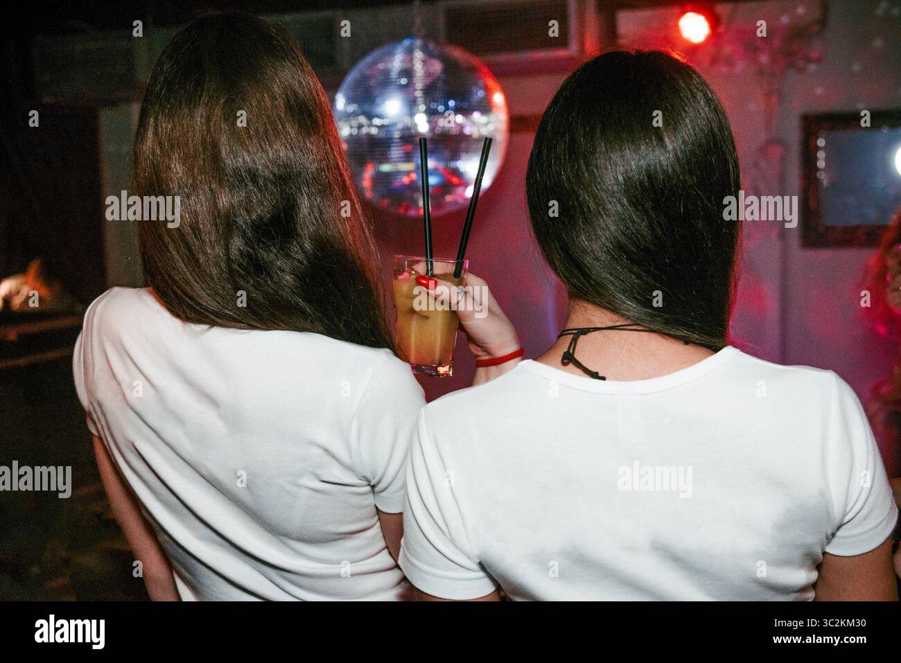 Deux jeunes femmes en t-shirts blancs se tiennent debout avec le dos et boivent des cocktails au bar du club lors de la fête Banque D'Images