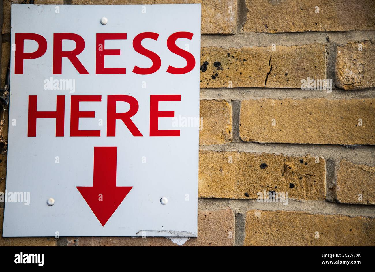 Grand signe 'PRESS HERE' en caractères rouges gras avec une flèche rouge pointant vers le bas, monté sur un mur de briques. Signalétique attirant l'attention, éventuellement pour l'emer Banque D'Images