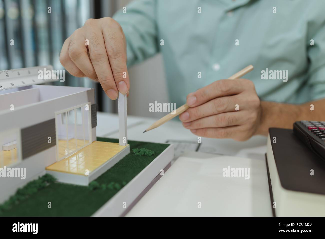 Un architecte dans une chemise vert clair travaillant sur un modèle de maison moderne à un bureau, assemblant soigneusement les pièces avec un crayon. Il se concentre sur la structure, Banque D'Images