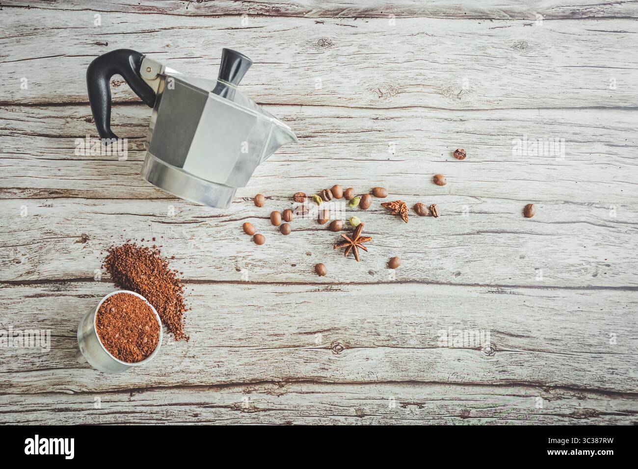 Pose plate de café ludique : le panier de moka-pot renverse de l'arabica moulu en forme de coeur, des haricots éparpillés, des épices et un brasseur incliné sur du bois rustique pâle, lumineux d Banque D'Images