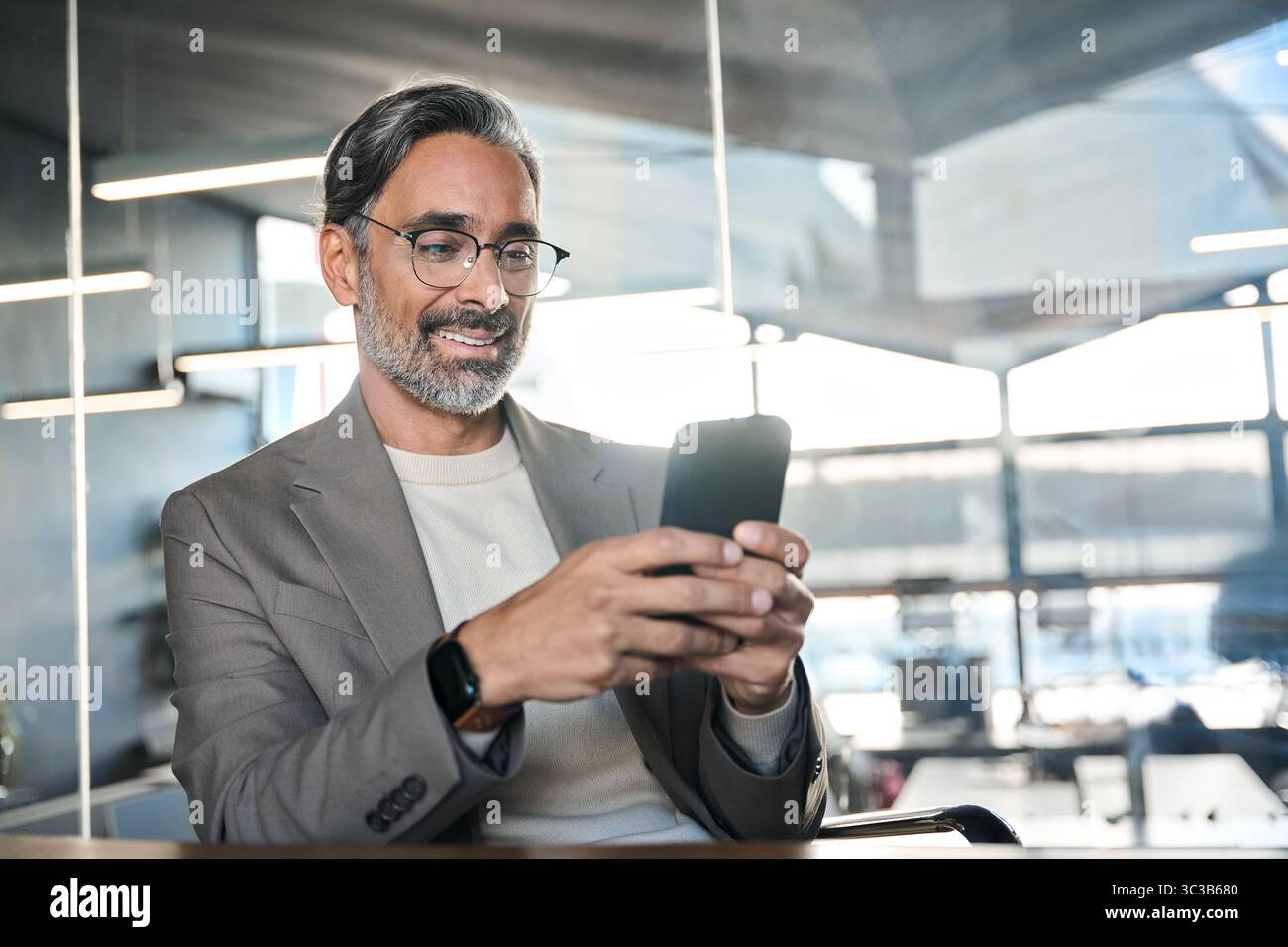 Homme d'affaires mûr souriant dirigeant assis à son bureau à l'aide d'un smartphone. Banque D'Images