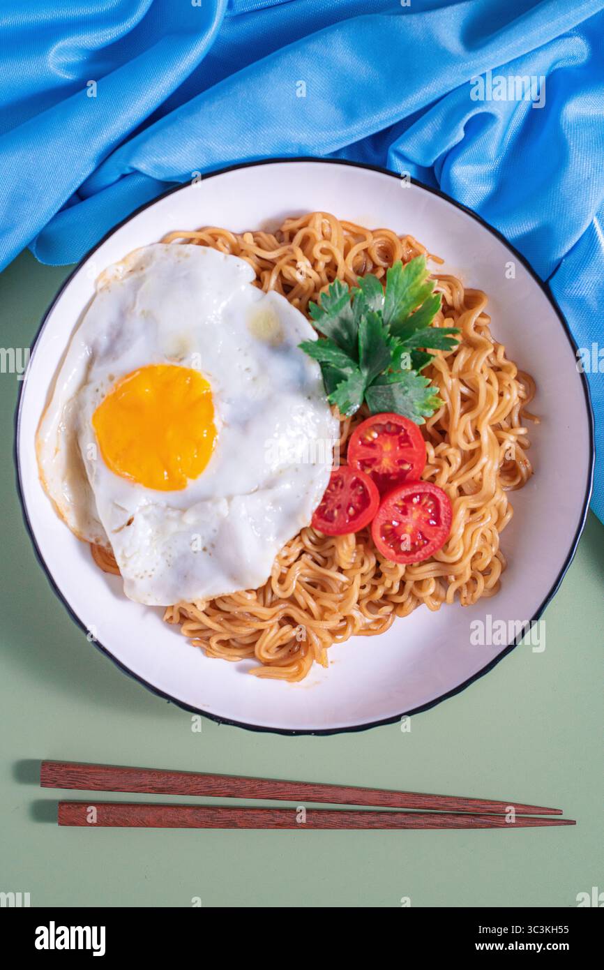 Nouilles instantanées cuites servies dans une assiette avec œuf ensoleillé, tranches de tomates et feuilles de céleri, complétées par des baguettes. Un comf savoureux et simple Banque D'Images