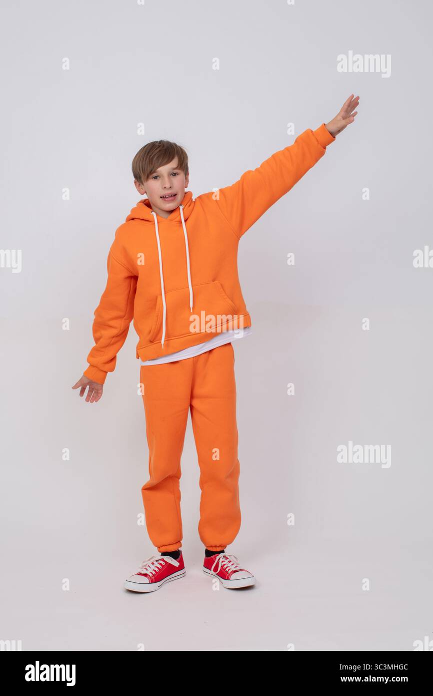 Un garçon portant un sweat-shirt orange et un pantalon orange leva la main et regarda la caméra. Le garçon porte des baskets rouges. Banque D'Images