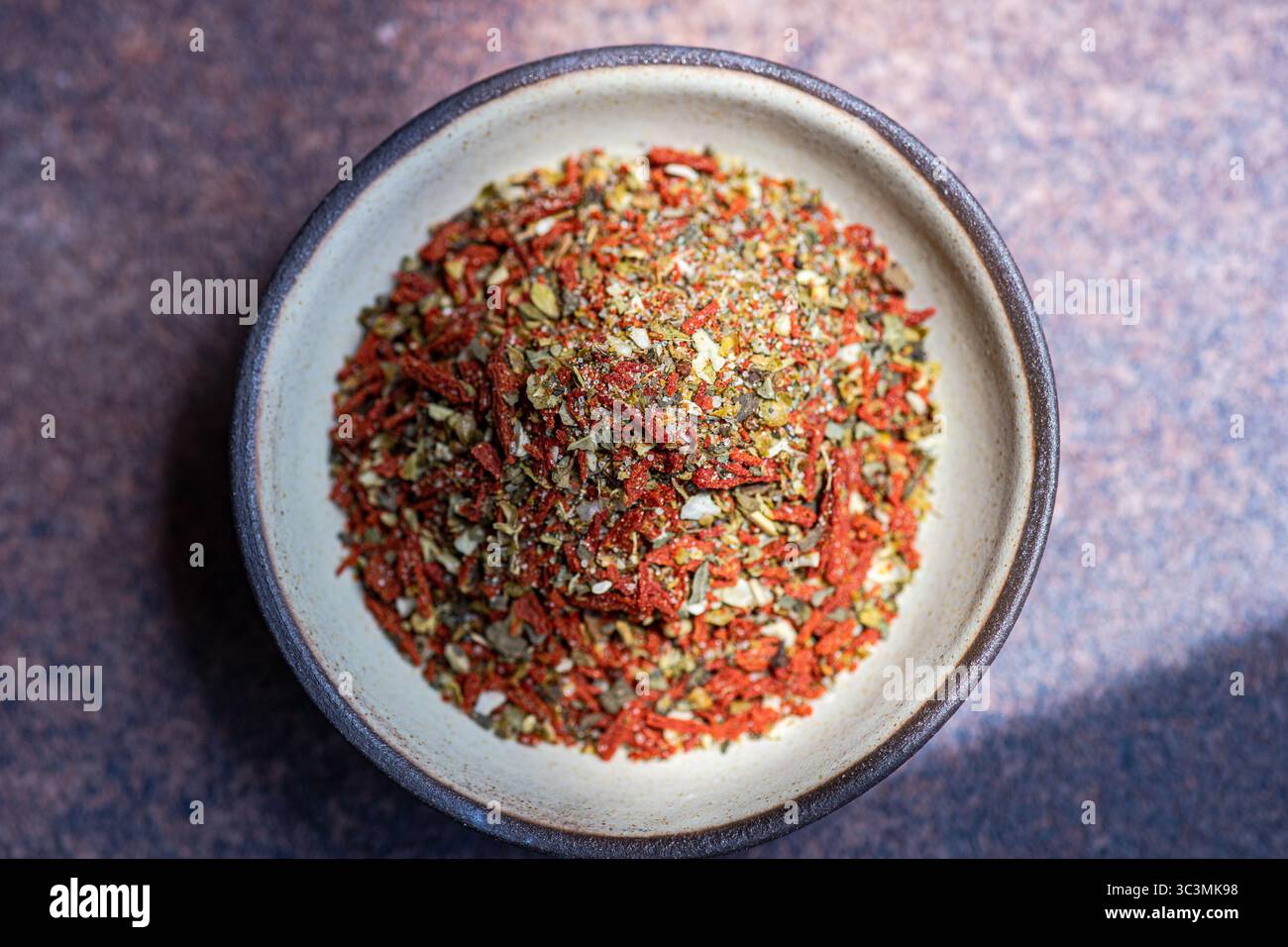 Vue de dessus d'un mélange d'épices vibrant contenant de la tomate et de l'olive, assaisonné de sel, exposé dans un bol en céramique beige contre un BA brun pierre texturé Banque D'Images
