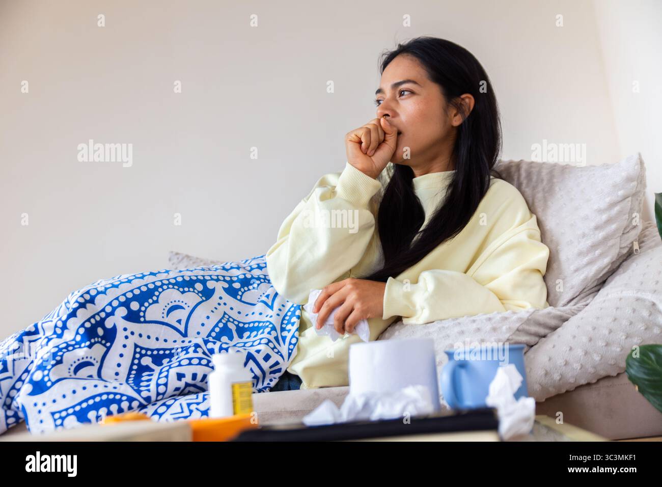 Une femme latine est assise sur un canapé confortable à la maison, paraissant malaise et réfléchie. Elle est enveloppée dans une couverture confortable, entourée de mouchoirs et de médicis Banque D'Images