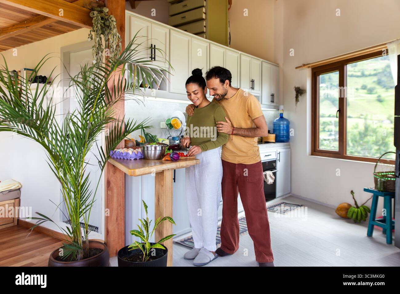 Un couple heureux profite d'un moment dans leur cuisine lumineuse, tranchant des légumes frais et préparant un repas ensemble. La salle bénéficie d'un éclairage naturel et d'une entrée Banque D'Images