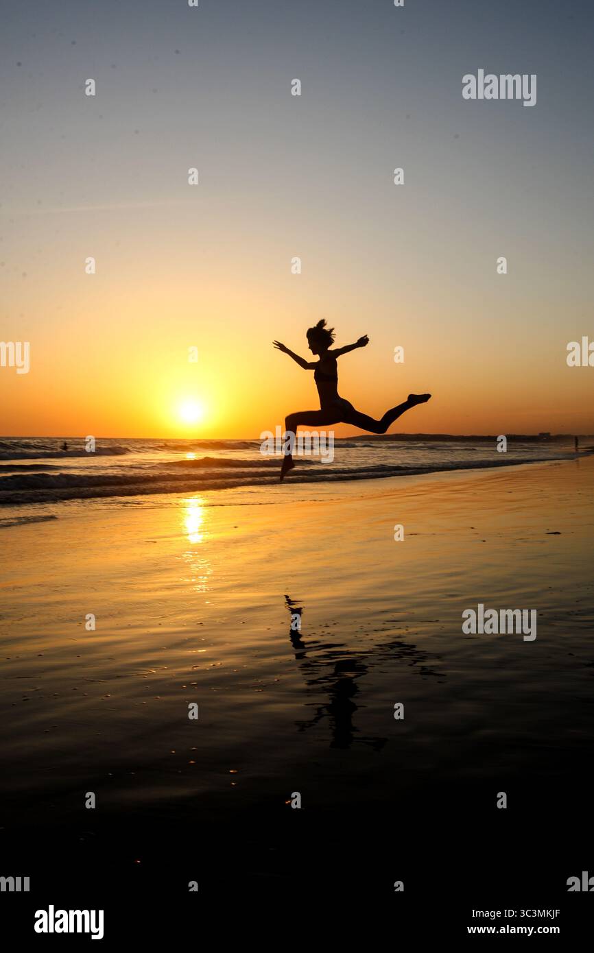 Silhouette de femme sportive en faisant un saut spectaculaire avec les jambes et les bras séparés sur la plage en face d'un beau coucher de soleil doré sur la mer. Banque D'Images