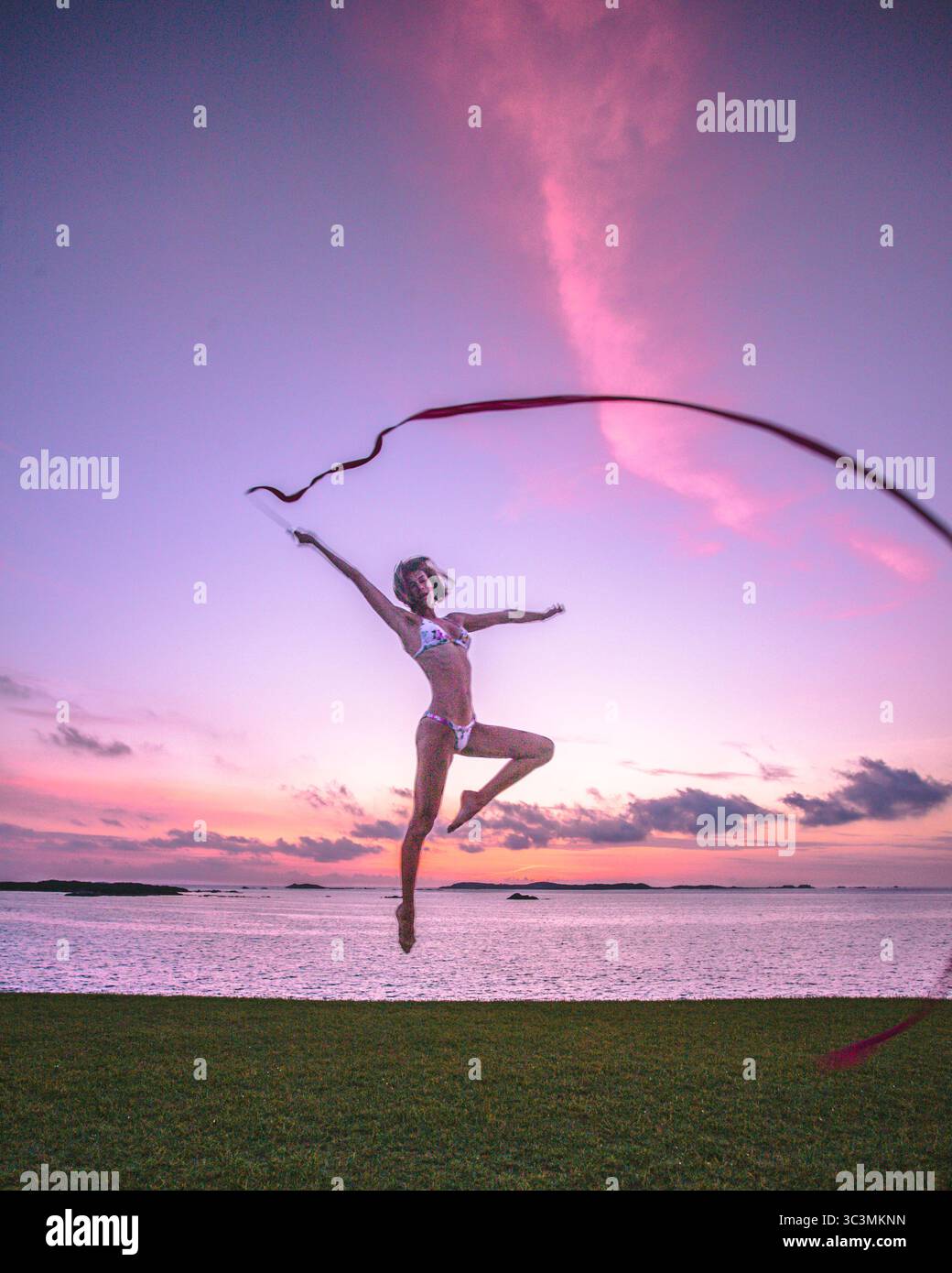 Femme sportive en bikini faisant un saut de danse avec ruban pour la gymnastique rythmique à la main devant un beau coucher de soleil sur la mer. Mode de vie actif Banque D'Images