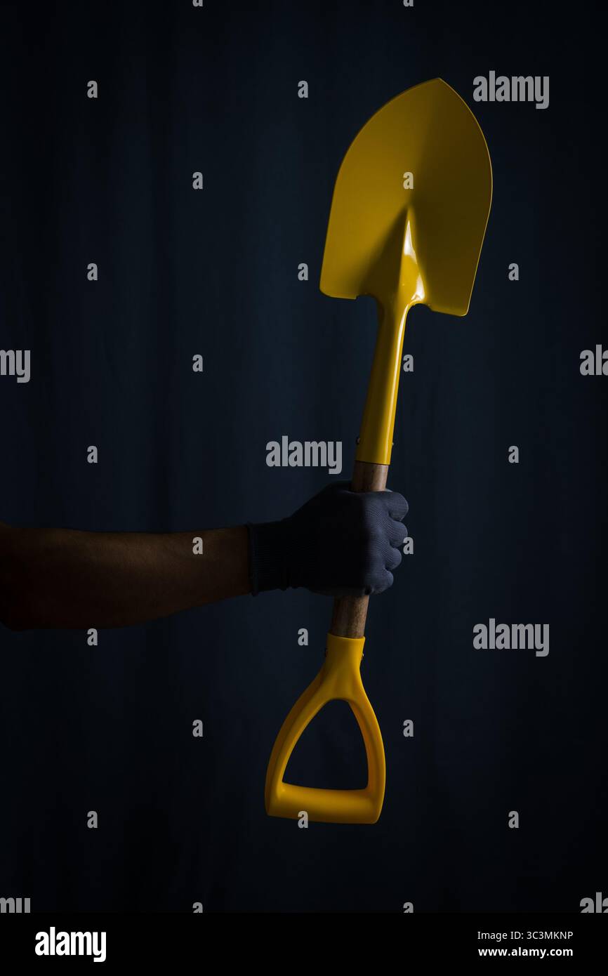 La main gantée de l'homme tient la pelle jaune sur fond sombre Banque D'Images