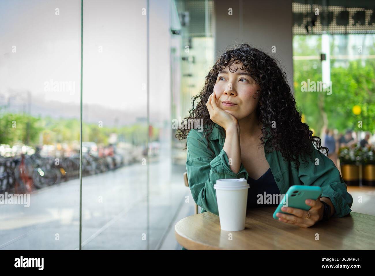 Jeune femme aux cheveux bouclés est assise dans un café moderne, tenant un smartphone et regardant dehors. Une tasse de café repose sur la table, entourée d'une urba calme Banque D'Images