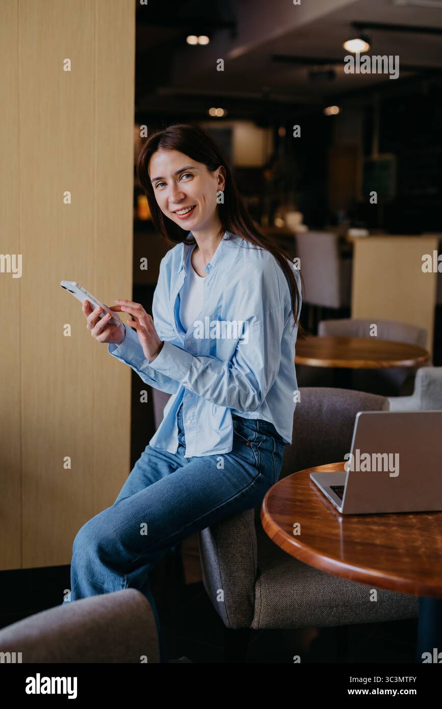 Une femme s'assoit confortablement dans un café confortable, s'engageant avec son smartphone et son ordinateur portable. Elle sourit et semble détendue, profitant de son environnement de travail, pendant ce temps Banque D'Images