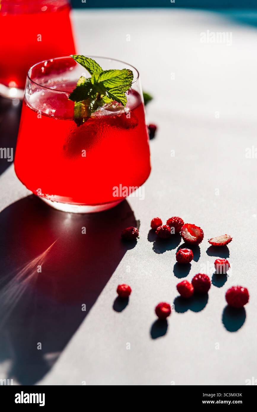 Verre rafraîchissant de fraises et de thé hibiscus est assis à la lumière directe du soleil, garni de menthe, entouré de baies fraîches dispersées sur un surf lumineux Banque D'Images
