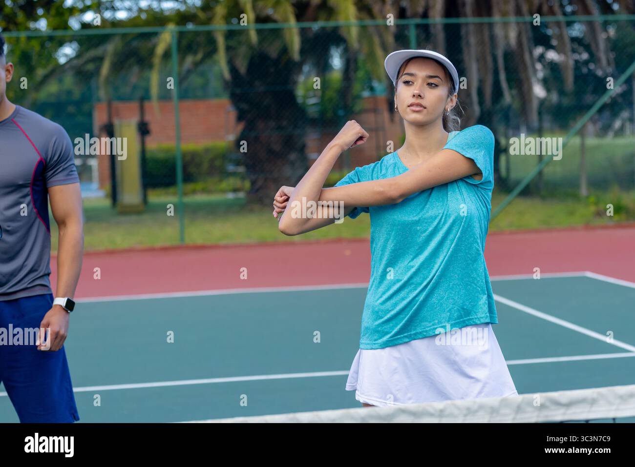 Partenaires de tennis réalisant un étirement de bras sur un court vert rouge par filet et clôture montrant une montre de sport Banque D'Images