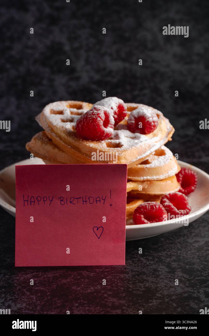 Une délicieuse pile de gaufres en forme de cœur, généreusement saupoudrées de sucre en poudre et de framboises fraîches, avec un joyeux anniversaire ! note dans le renoncement Banque D'Images