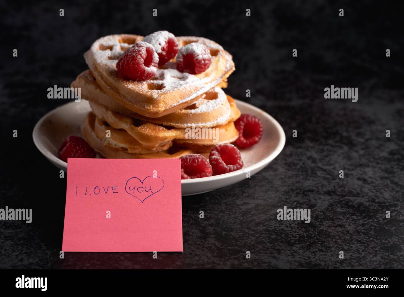 Une délicieuse pile de gaufres en forme de cœur, ornée de sucre en poudre et de framboises mûres, présentée à côté d'une note rose avec « I Love You » écrit Banque D'Images