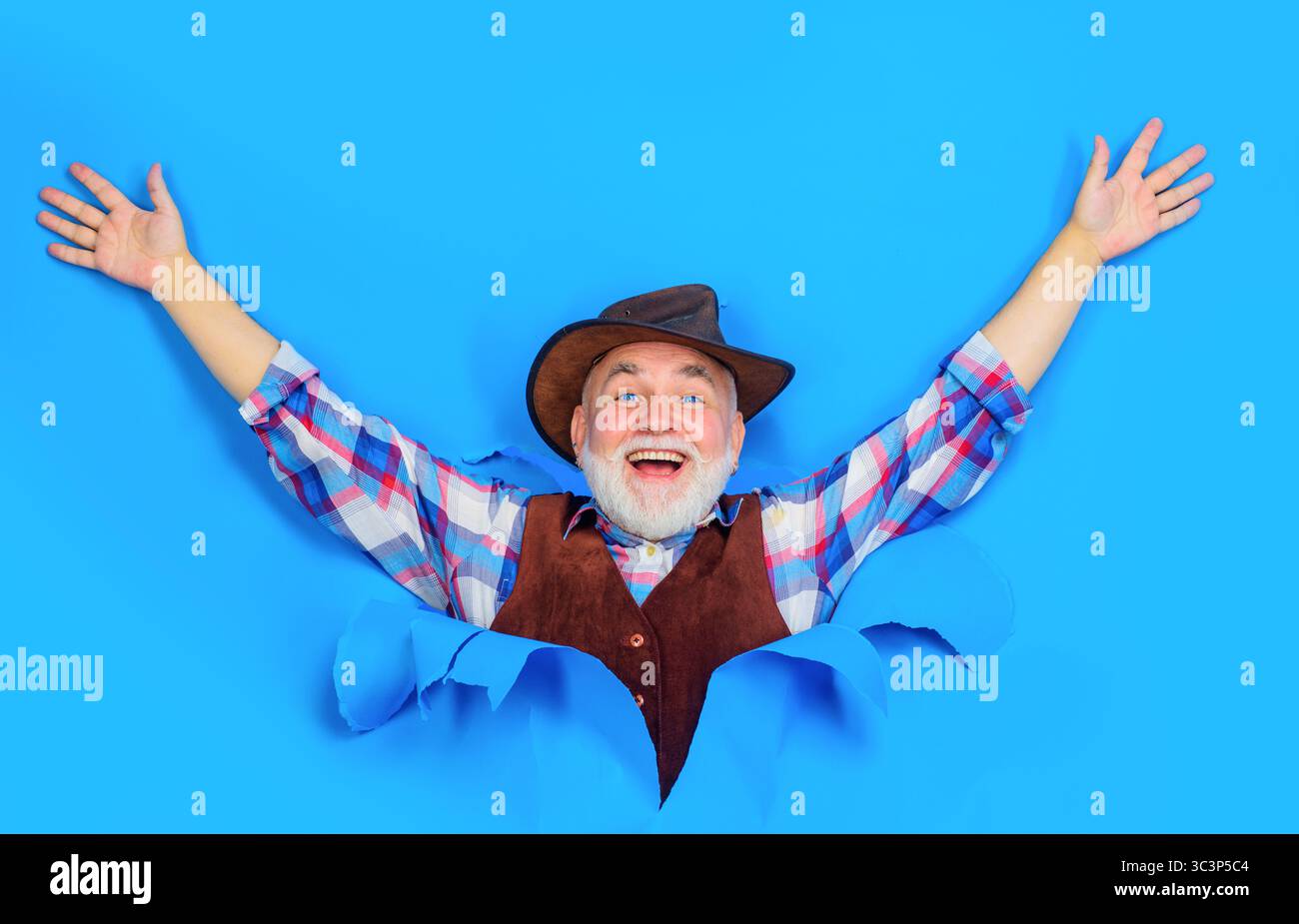 Joyeux vieil homme barbu en chemise à carreaux, gilet et chapeau de cow-boy en cuir regardant à travers le trou dans le papier. Beau senior en chapeau de cow-boy et chemise à carreaux Banque D'Images