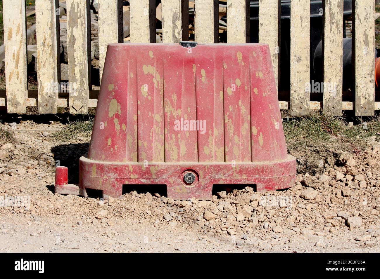 Une barricade en plastique rouge recouverte de saleté est partiellement encastrée dans du gravier brut sur un chantier de construction en bordure de route, signalant les dangers et séparant les zones de travail Banque D'Images