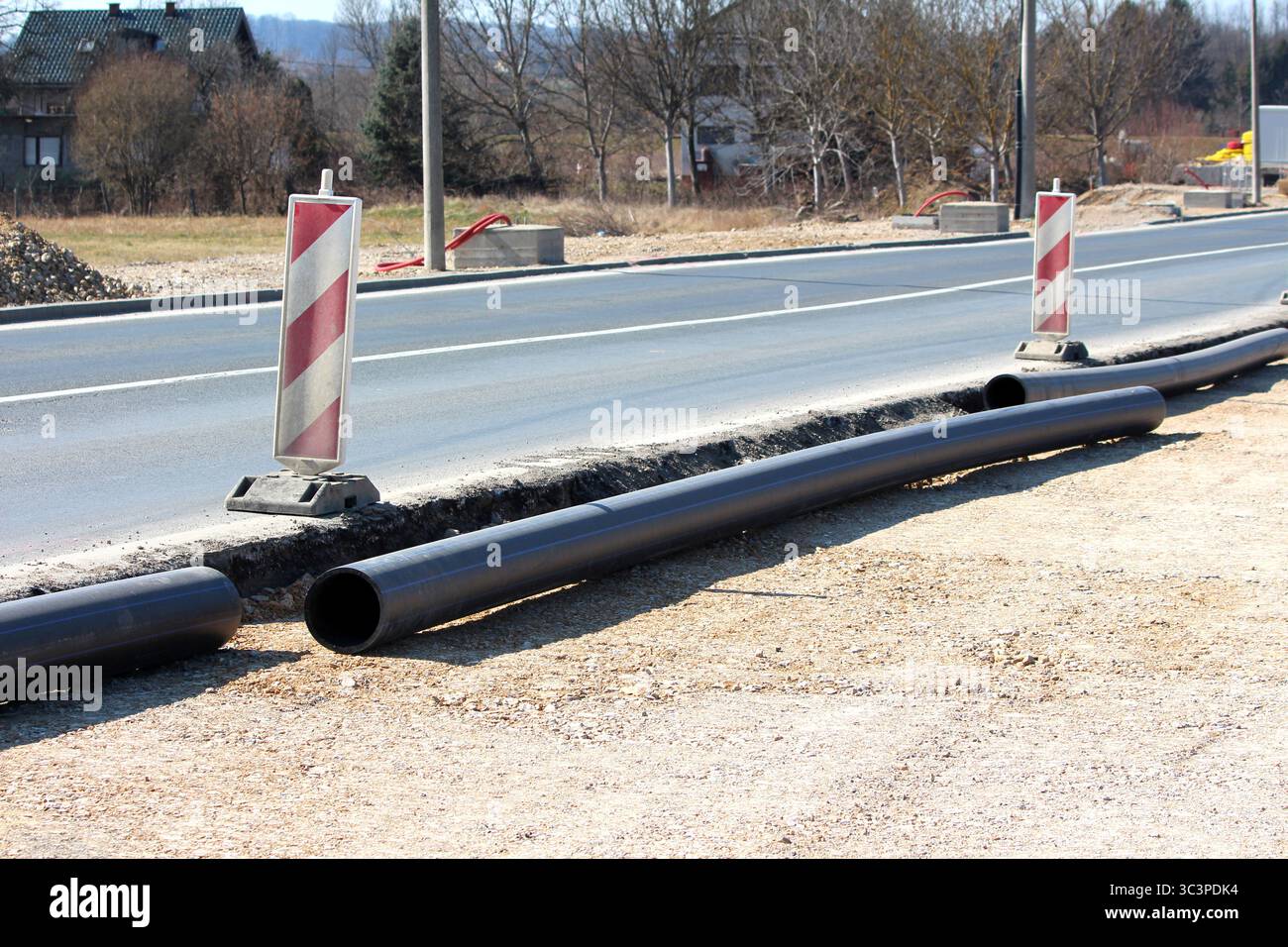 Des tuyaux de service publics en plastique noir sont disposés le long d'une route en cours d'entretien, marquée par des bornes de danger rayées, montrant des améliorations actives de l'infrastructure Banque D'Images