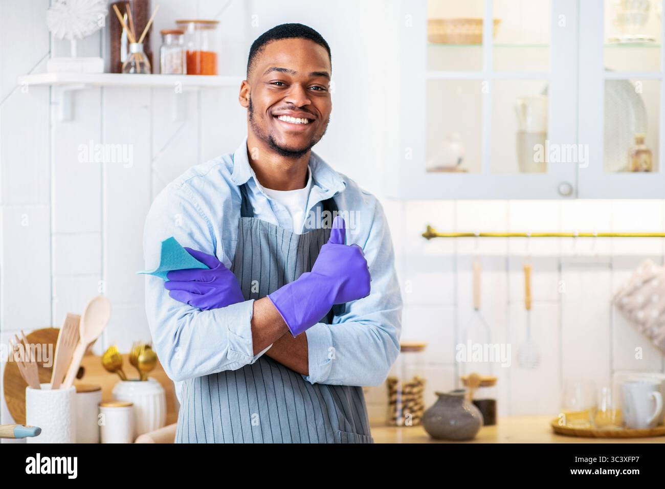 Beau jeune homme noir dans des gants en caoutchouc souriant à l'appareil photo et en montrant le pouce vers le haut dans la cuisine Banque D'Images