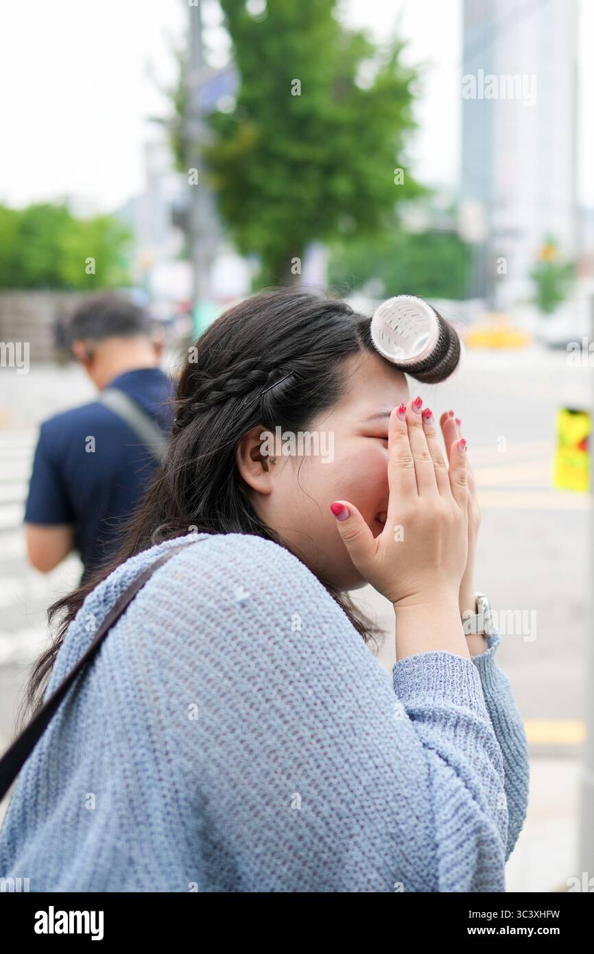 En juin, à Séoul, une coréenne d'une vingtaine d'années se tient debout sur un trottoir bondé de la ville, ajustant ses cheveux et se coiffant le visage avec les deux mains comme trafic Banque D'Images