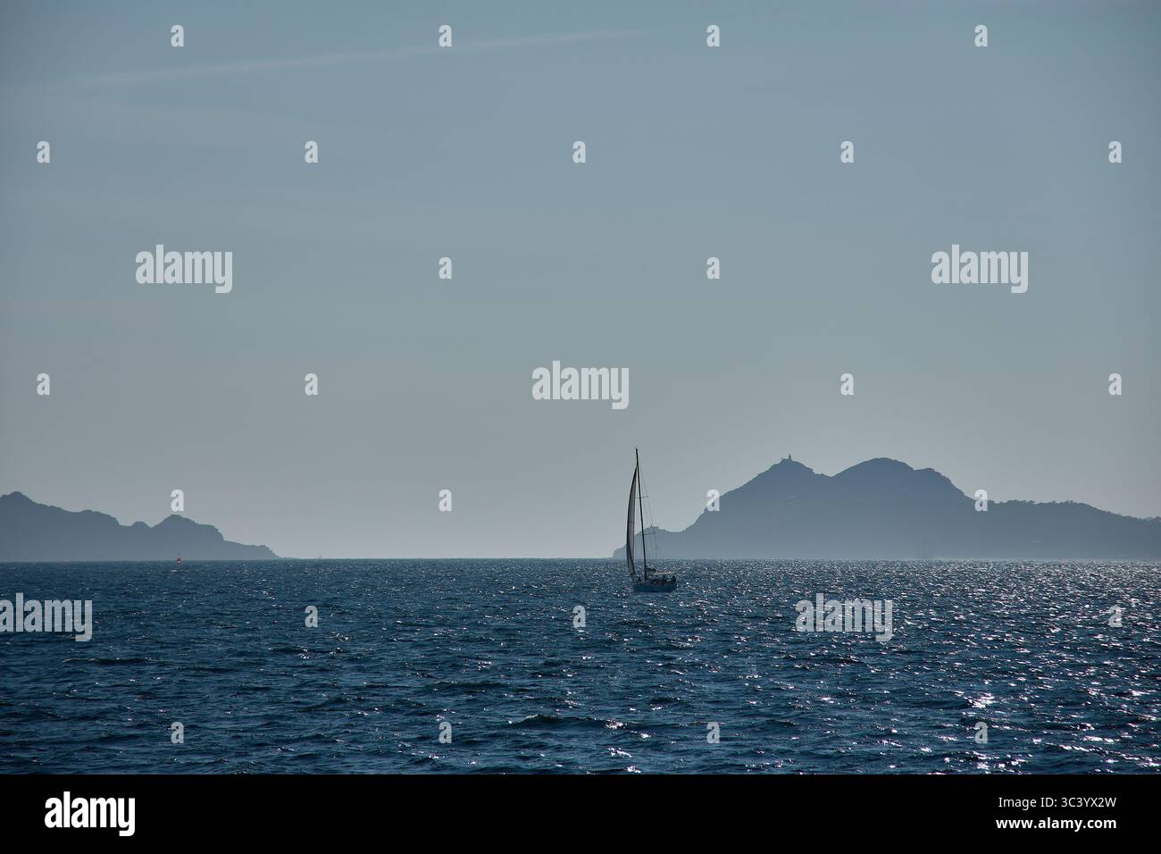 Un voilier navigue sereinement dans les eaux de l’estuaire de Vigo au coucher du soleil, avec la silhouette des îles Cíes Banque D'Images