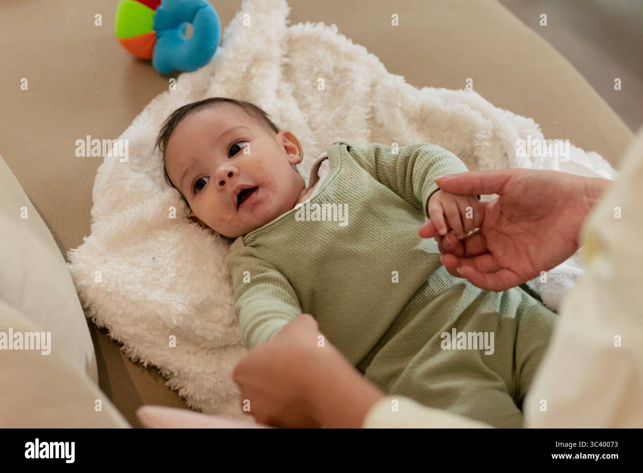 Mains des parents berçant bébé fille couché sur une couverture blanche en peluche à la maison avec jouet anneau multicolore Banque D'Images