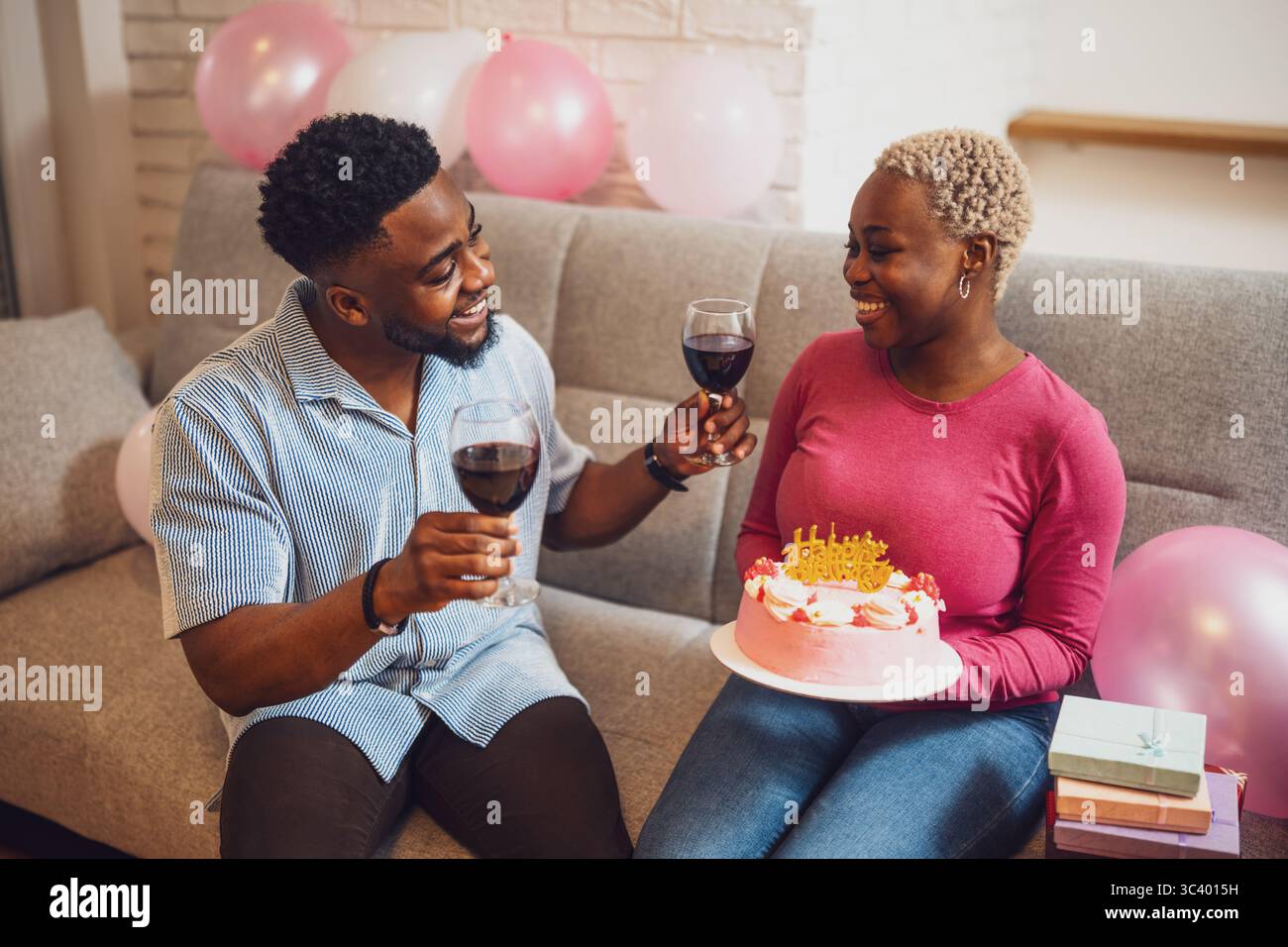 Heureux jeune couple célébrant l'anniversaire de la femme à la maison. La femme tient le gâteau et ils grillent avec du vin. Banque D'Images