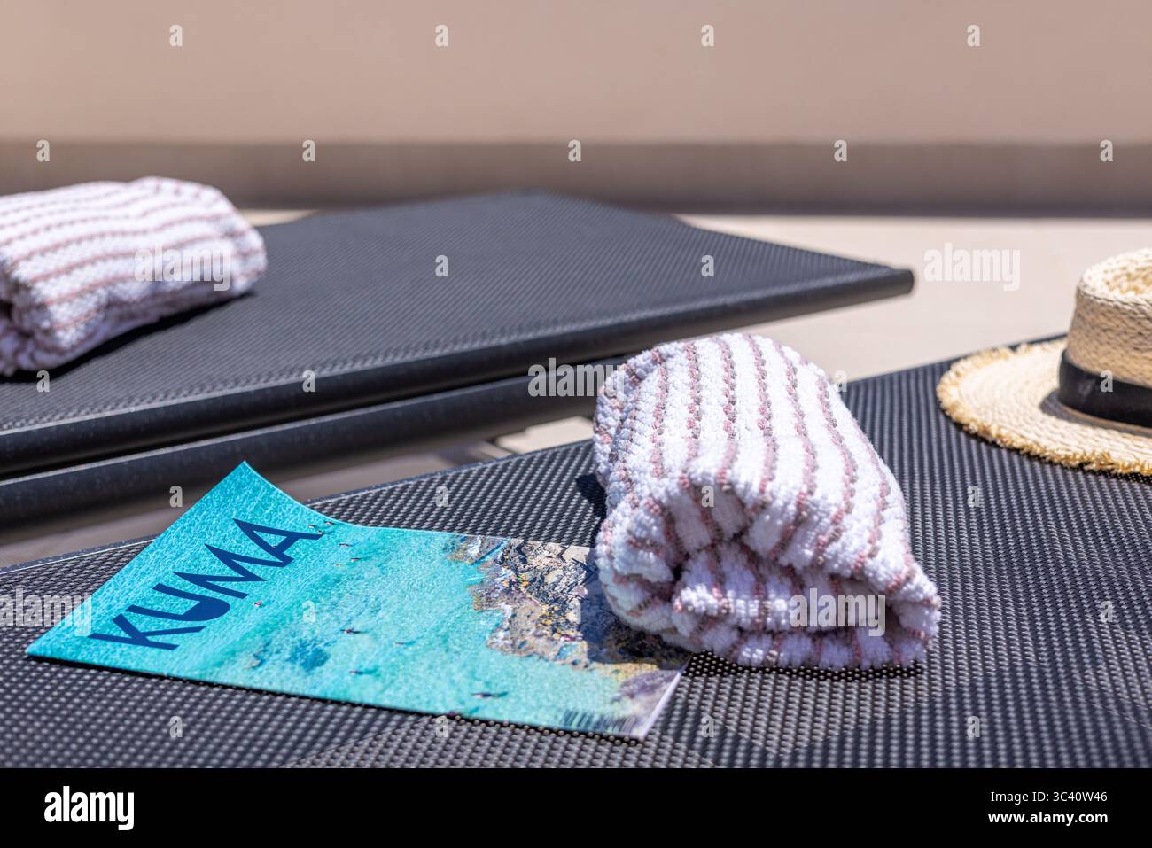 Gros plan sur les chaises longues au bord de la piscine avec une serviette rayée, un chapeau de paille et une brochure de voyage, évoquant une atmosphère de vacances estivales. Banque D'Images