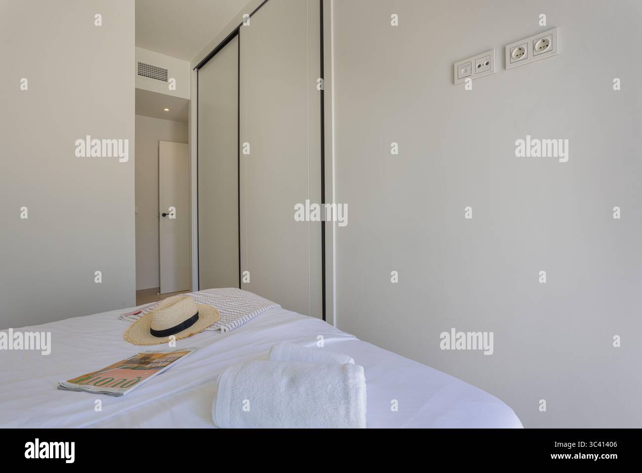Chambre minimaliste avec draps blancs, chapeau de paille et serviette sur le lit, grande armoire intégrée et accessoires modernes. Banque D'Images