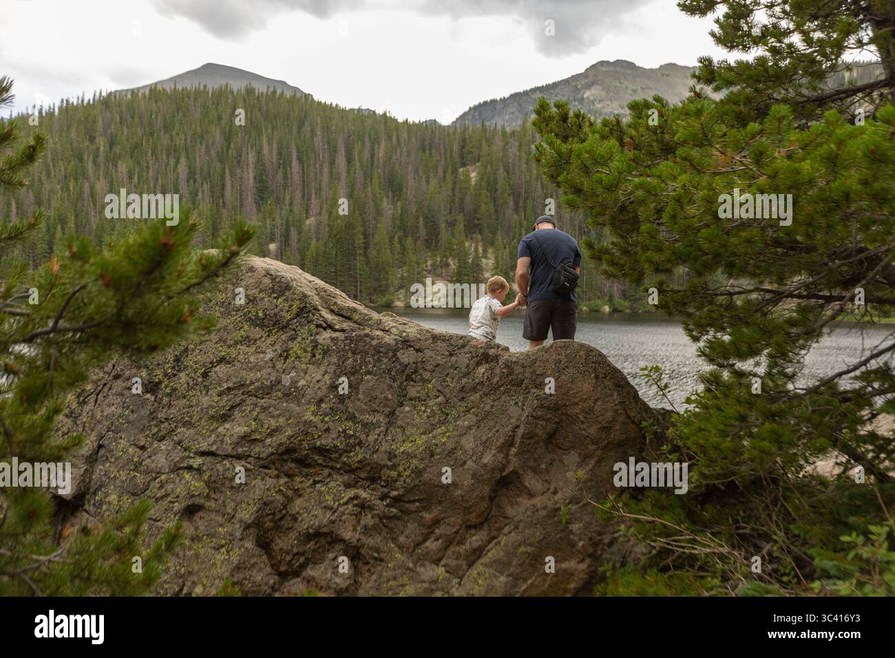 L'homme et l'enfant explorent les rochers du lac dans les montagnes couvertes de pins. Banque D'Images