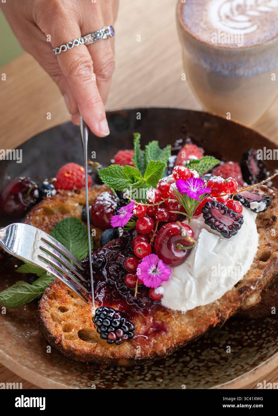 Gros plan d'une personne coupant une tranche de pain grillé garnie de baies fraîches et de crème à l'aide d'une fourchette et d'un couteau. Petit déjeuner sain ou brunch scène, fait maison. Banque D'Images