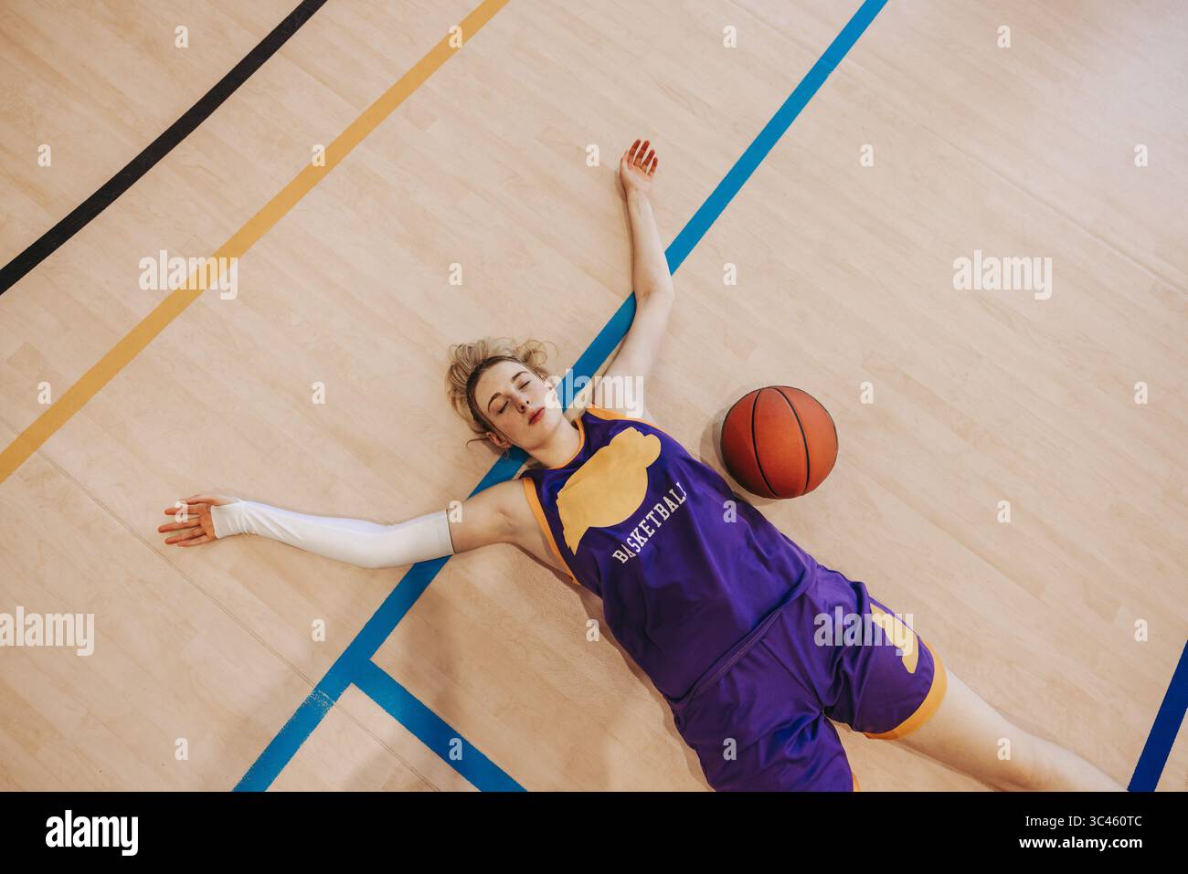 Une joueuse de basket-ball dans un uniforme violet couchée sur un terrain de gymnastique avec ses bras tendus et un ballon de basket à côté d'elle, transmettant un sentiment d'exhaustio Banque D'Images